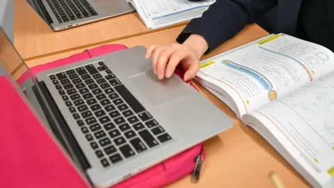A child wearing a navy jacket hovers their hand over a silver laptop, on top of a pink laptop case. They lean on a tan wooden table, with a maths textbook open next to them.