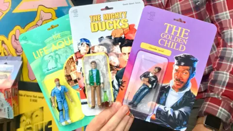 A man holding three home-made action figures. They look like real, professional packaged toys. The figures are all from movies - Eddie Murphy in The Golden Child, Bill Murray in The Life Aquatic, and Emilio Estevez in The Mighty Ducks.