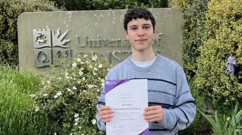 Daniel Virin wearing a grey striped jumper and holding a certificate for the Innovation Outstanding PLUS awards. He is standing in front of a University of Bristol sign, which is nestled among bushes and white flowers. He has dark curly hair and is smiling at the camera.